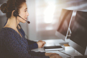 Two busineswomen have conversations with the clients by headsets, while sitting at the desk in a sunny modern office. Diverse people group in a call center. Telemarketing and customer service